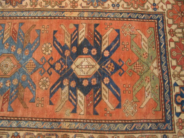 Geometric rug detail showing a rust-colored field with stylized indigo and sage-green figures and wave borders.