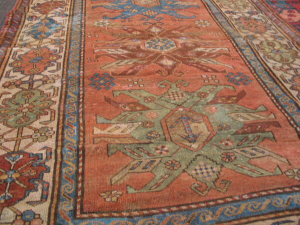 Geometric rug with rust field and complex borders featuring blue, tan, and olive green patterns.