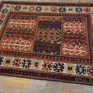 Tribal rug featuring geometric patterns in reddish-orange, navy, and cream on gray wood flooring.
