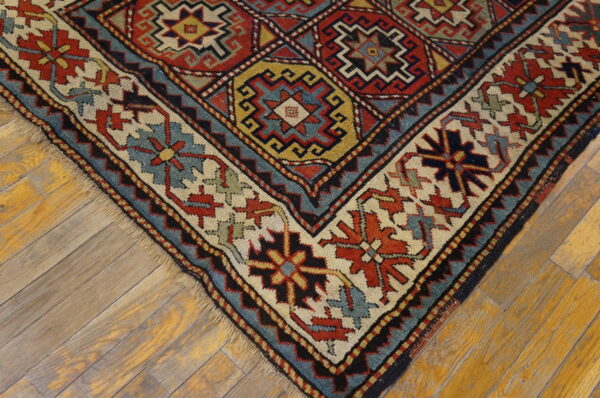 Low-pile rug corner showing red, cream, blue, and yellow geometric patterns on weathered wood planks.