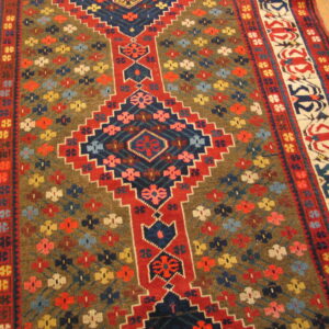 Low-pile rug with red, navy, and brown geometric patterns and floral accents resting on a wood floor.