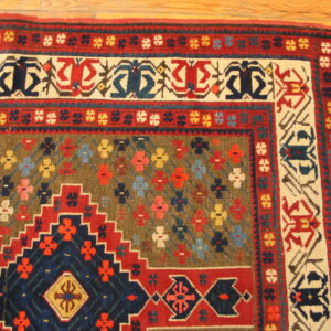 Detailed corner of a low-pile rug in red, navy, and olive geometric patterns, sitting on hardwood floor planks.