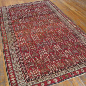 A rectangular, deep red rug with intricate geometric patterns and multiple borders rests on a wooden floor.