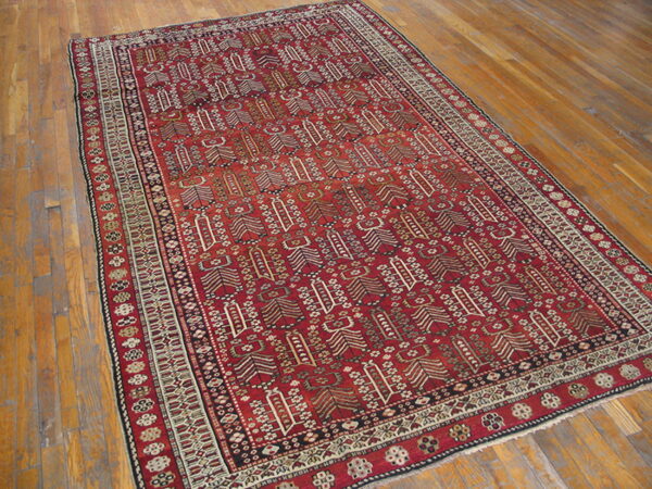 A rectangular, deep red rug with intricate geometric patterns and multiple borders rests on a wooden floor.