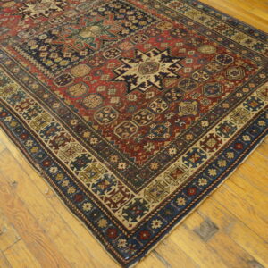 A geometric low-pile rug in red, navy, and tan colors is displayed on rustic plank wood flooring.