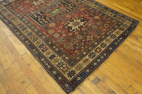 A geometric low-pile rug in red, navy, and tan colors is displayed on rustic plank wood flooring.