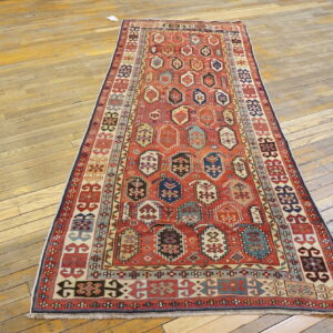 Long runner rug features a red geometric field with multi-colored guls over worn wood planks.