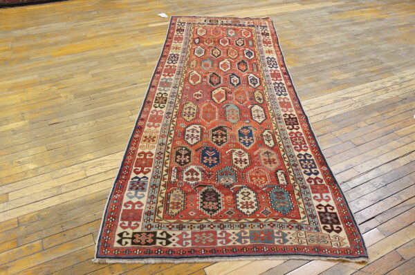 Long runner rug features a red geometric field with multi-colored guls over worn wood planks.