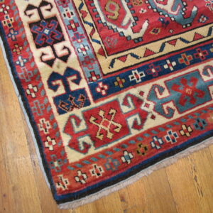 Close-up of a red, cream, and blue geometric patterned rug corner on light brown wood flooring.