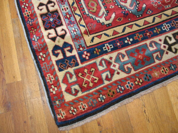 Close-up of a red, cream, and blue geometric patterned rug corner on light brown wood flooring.