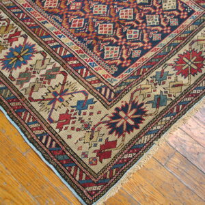 A detailed corner of a dense, patterned rug displaying red, navy, and tan colors on worn wooden planks.
