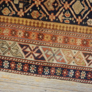 Geometric rug close-up displays repeating bands of navy, brown, rust, and tan designs over light wood.