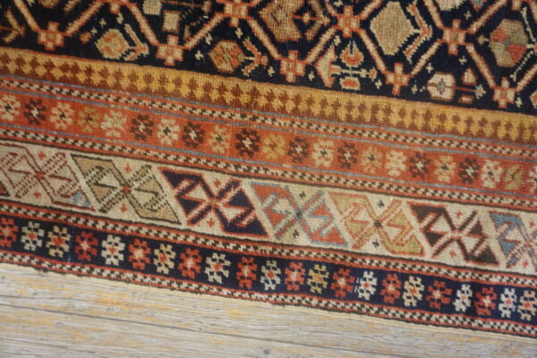 Geometric rug close-up displays repeating bands of navy, brown, rust, and tan designs over light wood.