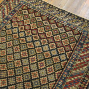 A low-pile rug with a dark field, colorful geometric lattice design, and striped borders rests on wood planks.