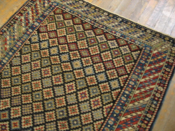 A low-pile rug with a dark field, colorful geometric lattice design, and striped borders rests on wood planks.