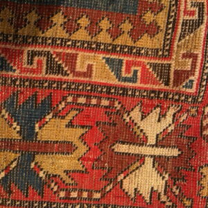 Geometric rug pattern close-up showing red, gold, dark blue, and brown colors next to a measuring tape.