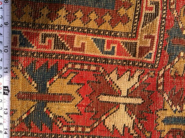 Geometric rug pattern close-up showing red, gold, dark blue, and brown colors next to a measuring tape.