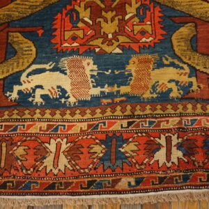 Detail of a low-pile rug in red, blue, and gold with lions and geometric bands, resting on wood.