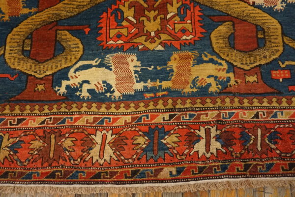 Detail of a low-pile rug in red, blue, and gold with lions and geometric bands, resting on wood.