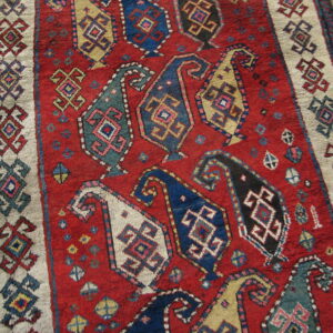 Textured red rug displays repeating colorful boteh patterns and a thick beige geometric border on wood flooring.