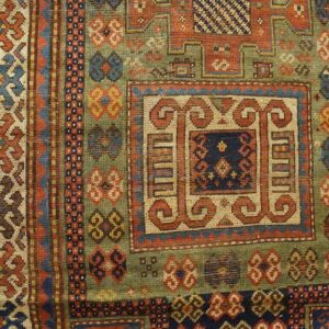 Detailed view of a low-pile rug with geometric patterns in olive green, rust, and blue on wood planks.
