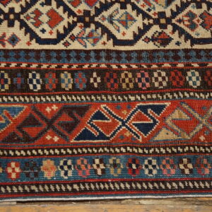 Low-pile geometric rug in red, navy, cream, and brown with intricate borders visible over wooden flooring.