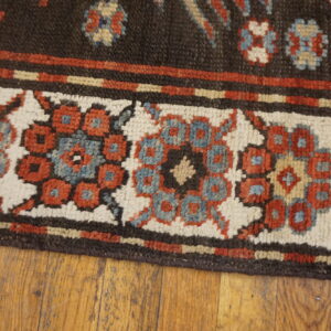 Dark brown rug edge with a patterned cream, red, and blue border lying on wood flooring.