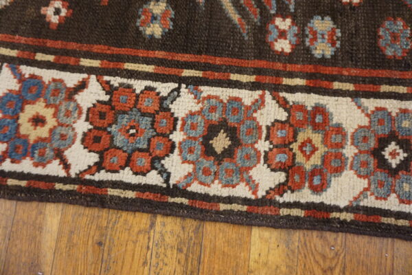 Dark brown rug edge with a patterned cream, red, and blue border lying on wood flooring.