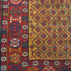 A tightly woven rug with red and blue borders and a gold diamond field rests on light hardwood flooring.