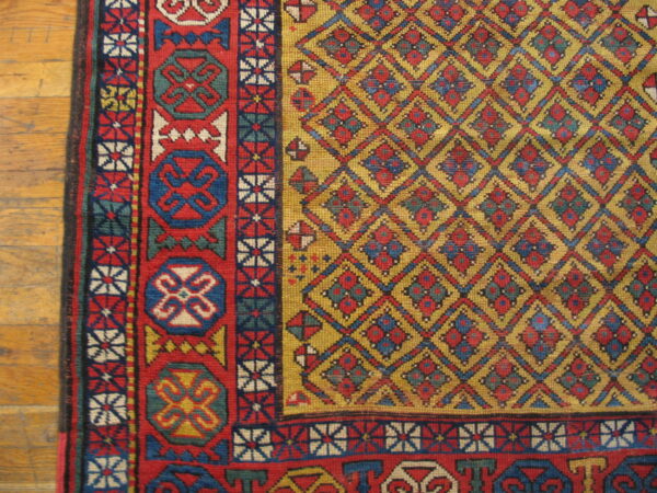 A tightly woven rug with red and blue borders and a gold diamond field rests on light hardwood flooring.