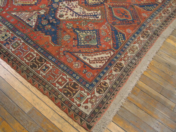 Red and dark blue geometric rug with multiple patterned borders and fringe rests on wood floorboards.