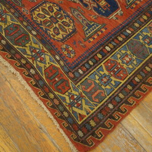 Low-pile rug corner in red, blue, gold, and brown geometric patterns rests on distressed wood planks.