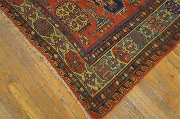 Low-pile rug corner in red, blue, gold, and brown geometric patterns rests on distressed wood planks.