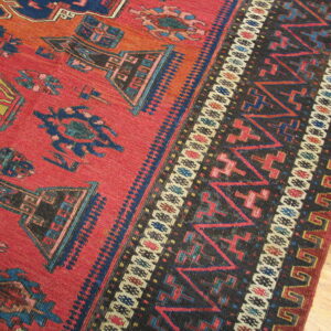 A low-pile red rug displays geometric tribal motifs and a complex dark border on wood flooring.