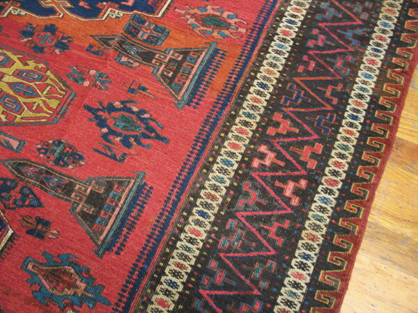 A low-pile red rug displays geometric tribal motifs and a complex dark border on wood flooring.