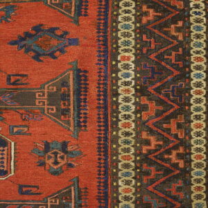 Low-pile rug detail showing a rust-red field and complex geometric borders in charcoal, blue, and tan.