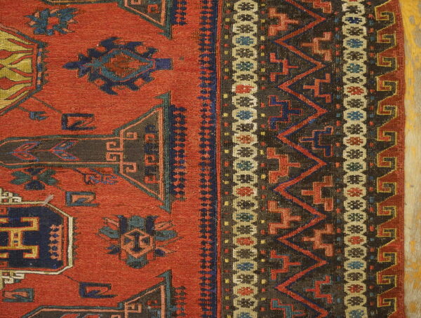 Low-pile rug detail showing a rust-red field and complex geometric borders in charcoal, blue, and tan.