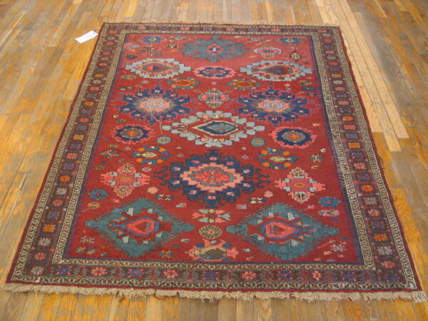 Red low-pile rug with geometric patterns in blue, teal, and pink rests on light plank wood flooring.