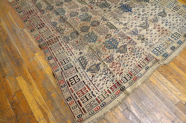 Beige rug with multicolored geometric motifs and stylized trees rests on worn wood flooring.