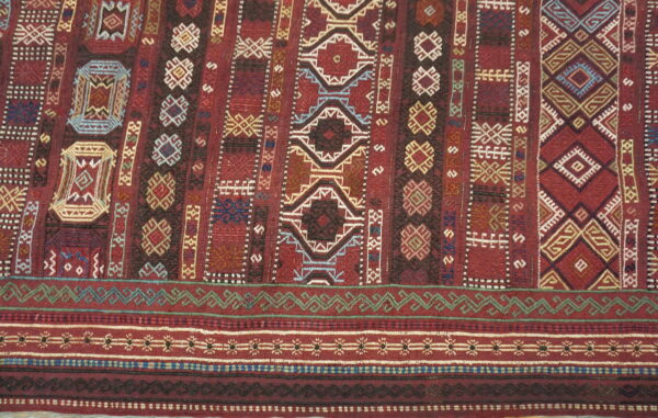 Flat-woven rug in deep red, patterned with complex geometric motifs and border bands in blue, green, and cream.