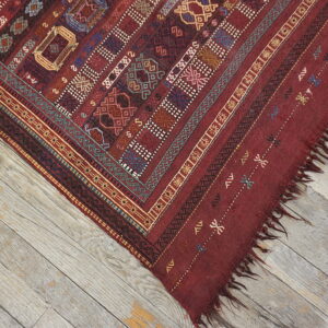 The corner of a red flatweave rug with detailed multicolor patterns lies on rustic gray wood planks.