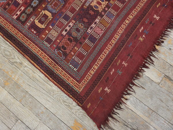 The corner of a red flatweave rug with detailed multicolor patterns lies on rustic gray wood planks.