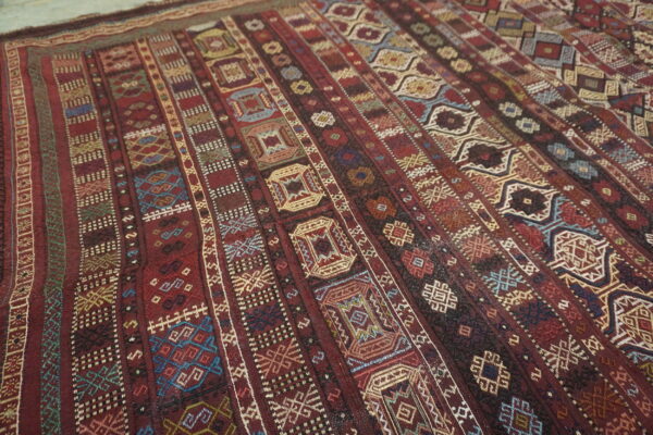 Flatweave textile displaying complex geometric patterns in vertical bands of deep red, gold, and blue.