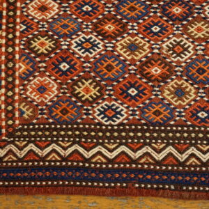 Geometric flatweave rug corner shows diamond motifs and intricate zigzag borders in rust, navy, and brown on wood flooring.