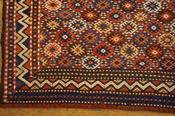 Geometric flatweave rug corner shows diamond motifs and intricate zigzag borders in rust, navy, and brown on wood flooring.