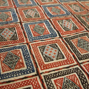 Textured rug surface displays a grid of red, blue, and black geometric designs on a cream ground.