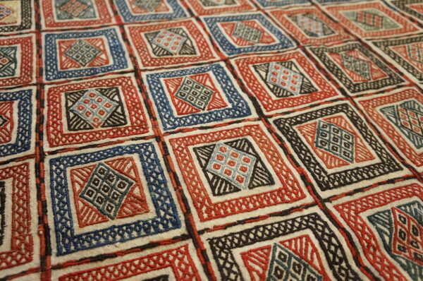 Textured rug surface displays a grid of red, blue, and black geometric designs on a cream ground.