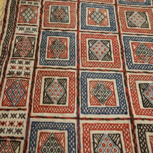 Low-pile rug features a grid of red and blue squares with geometric motifs resting on hardwood planks.