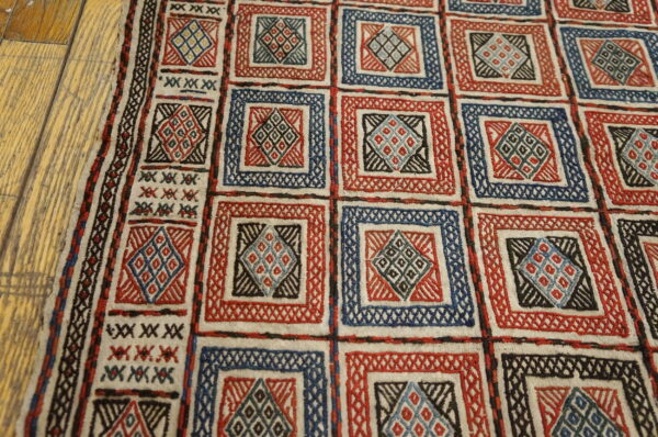 Low-pile rug features a grid of red and blue squares with geometric motifs resting on hardwood planks.