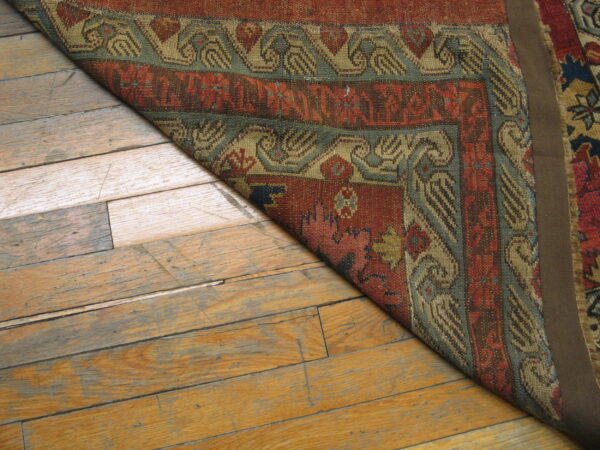 A rust and teal patterned rug is folded back on worn, wide-plank wood flooring.